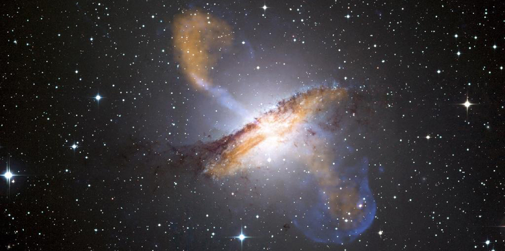 A photo of deep space showing a lot of bright spots on a black background. In the center is a huge ring of brownish gas that encircles an interior that is glowing white. Along the axis of the ring more brownish bluish gas is spewing out into the surrounding space.