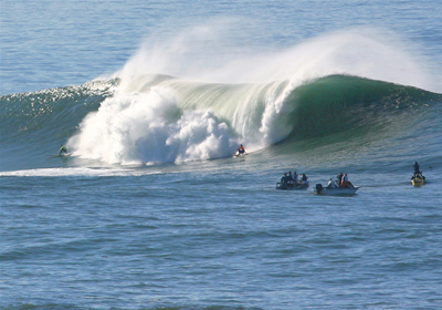 There is a high tidal wave of about 10 feet high in a sea. Three boats carrying three four persons each are ahead of the wave, which is coming toward them.